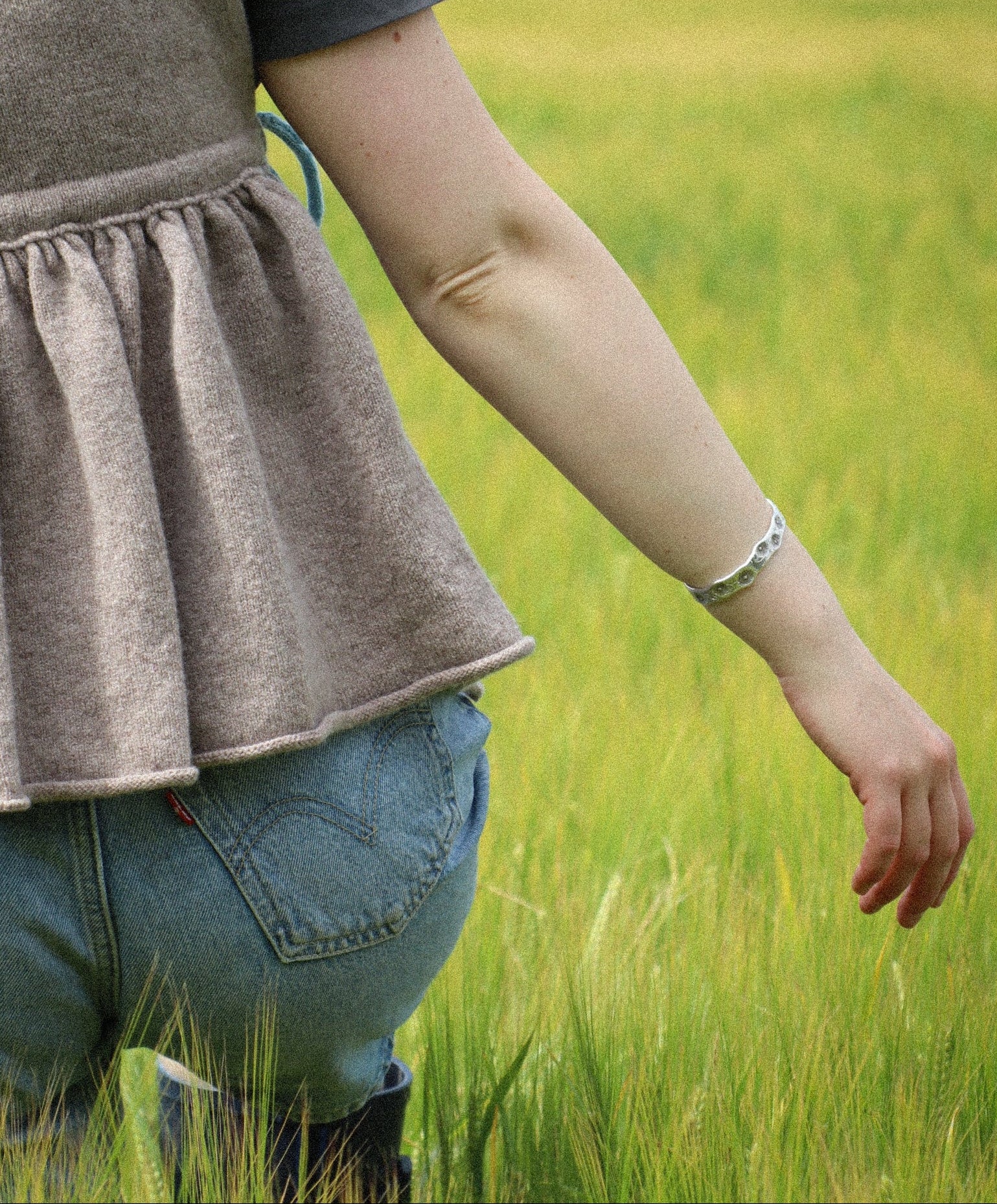 A person standing in a green field, shown from the back wearing a taupe pleated vest over a dark gray t-shirt and blue jeans.